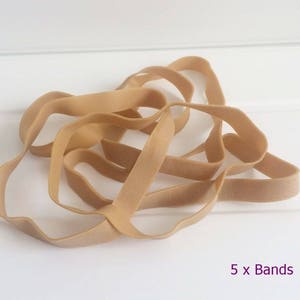 May include: A close-up of five tan-coloured rubber bands, looped and intertwined. The bands are arranged in a tangled pattern. The text "5 x Bands" is visible in the lower right corner.