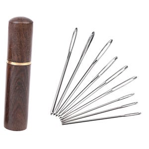 May include: A collection of sewing needles of different lengths, arranged in a fan shape, beside a dark brown wooden needle case with a gold band. The needles are made of shiny, silver-coloured metal. The case is cylindrical with a rounded top.