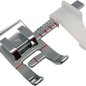 May include: A silver sewing machine foot with red markings and a white plastic guide. The foot has a flat base with two raised bars and a curved front edge. The guide extends upwards and to the right.