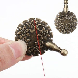 May include: A vintage-style, gold-toned thread dispenser with a floral design. The dispenser has a small loop at the top for hanging and a small handle on the side. A red thread is being pulled through the dispenser.