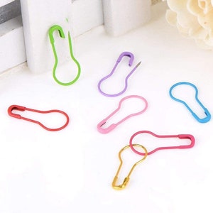 May include: A set of seven colorful safety pins in various colors, including green, purple, pink, red, blue, and gold. The pins are all in the shape of a light bulb.