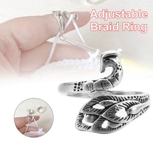 May include: Silver adjustable braid ring with a decorative design. The ring is open and can be adjusted to fit different finger sizes. The ring is designed to hold yarn while crocheting.