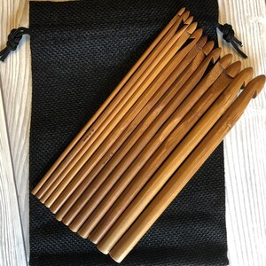 以下が含まれることがあります： 様々なサイズの木製かぎ針編みフックのセット。黒い巾着袋の上に置かれています。フックは明るい茶色で、竹でできているようです。バッグは、質感のある黒い生地で作られています。