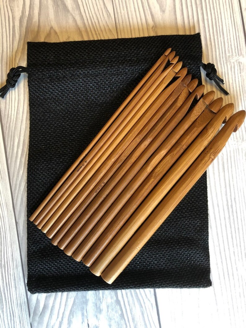 以下が含まれることがあります： 様々なサイズの木製かぎ針編みフックのセット。黒い巾着袋の上に置かれています。フックは明るい茶色で、竹でできているようです。バッグは、質感のある黒い生地で作られています。
