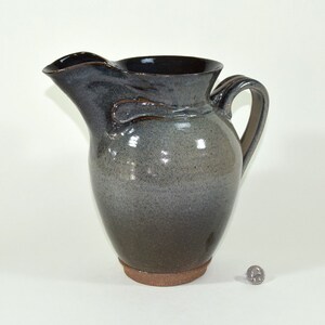 May include: A handmade ceramic pitcher with a dark gray and brown glaze. The pitcher has a curved spout, a handle, and a textured base. The overall shape is rounded, with a smooth, glossy finish.