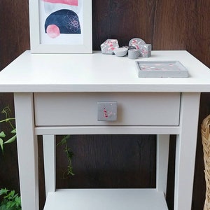 Può includere: Tavolino bianco con un cassetto e uno scaffale inferiore. Il cassetto ha una manopola macchiata di grigio e rosa. Il piano del tavolo ha un vassoio macchiato di grigio e rosa e diversi piccoli oggetti decorativi macchiati di grigio e rosa.