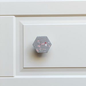 Puede incluir: Un pomo hexagonal para armario hecho de hormigón gris con motas rosas y rojas.