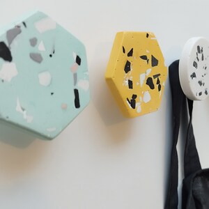 K&ouml;nnte beinhalten: Vier sechseckige Haken mit einem Terrazzo-Muster in Gr&uuml;n-, Gelb-, Wei&szlig;- und Graut&ouml;nen. Die Haken sind an einer wei&szlig;en Wand montiert und ein schwarzer Gurt h&auml;ngt an einem der Haken.