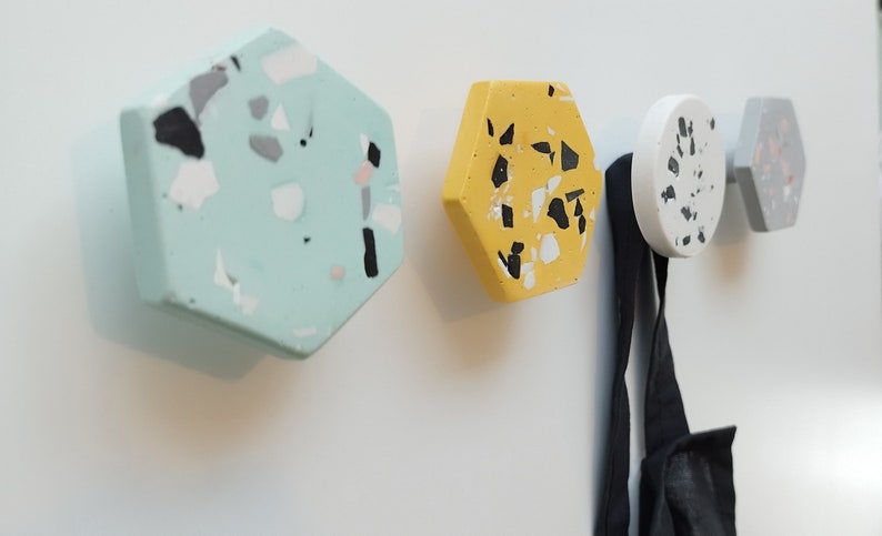 K&ouml;nnte beinhalten: Vier sechseckige Haken mit einem Terrazzo-Muster in Gr&uuml;n-, Gelb-, Wei&szlig;- und Graut&ouml;nen. Die Haken sind an einer wei&szlig;en Wand montiert und ein schwarzer Gurt h&auml;ngt an einem der Haken.