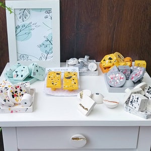 K&ouml;nnte beinhalten: Sortiment an bunten, gesprenkelten und gemusterten Betonschubladen- und -kn&auml;ufen. Die Kn&ouml;pfe haben verschiedene Formen, darunter rund, quadratisch und rechteckig. Die Griffe sind in verschiedenen Farben, darunter Gelb, Grau und Rosa. Die Kn&ouml;pfe und Griffe sind auf einer wei&szlig;en Oberfl&auml;che mit einem wei&szlig;en Rahmen und einer wei&szlig;en Schublade angeordnet.