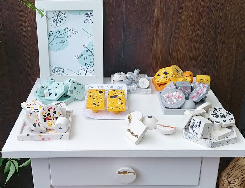 K&ouml;nnte beinhalten: Sortiment an bunten, gesprenkelten und gemusterten Betonschubladen- und -kn&auml;ufen. Die Kn&ouml;pfe haben verschiedene Formen, darunter rund, quadratisch und rechteckig. Die Griffe sind in verschiedenen Farben, darunter Gelb, Grau und Rosa. Die Kn&ouml;pfe und Griffe sind auf einer wei&szlig;en Oberfl&auml;che mit einem wei&szlig;en Rahmen und einer wei&szlig;en Schublade angeordnet.