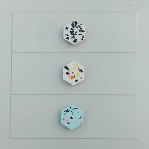 K&ouml;nnte beinhalten: Drei sechseckige Schrankkn&ouml;pfe mit Terrazzo-Design. Der obere Knopf ist schwarz-wei&szlig;, der mittlere Knopf ist wei&szlig; mit orangefarbenen, gelben und schwarzen Flecken und der untere Knopf ist hellblau mit rosa, wei&szlig;en und schwarzen Flecken.