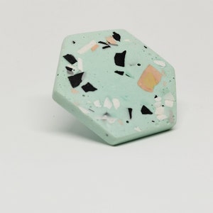 K&ouml;nnte beinhalten: Ein sechseckiger Untersetzer mit einer hellgr&uuml;nen Basis und einem gesprenkelten Muster aus Schwarz, Wei&szlig; und Pfirsich.