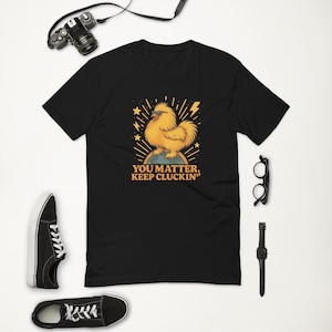 Könnte beinhalten: Schwarzes T-Shirt mit einem Cartoon-Huhn mit Sonnenbrille, das auf einem Globus steht, mit dem Text "YOU MATTER. KEEP CLUCKIN". Das Design umfasst Sterne und Blitze. Das T-Shirt ist von einer Kamera, Schuhen, einer Brille und einer Uhr umgeben.