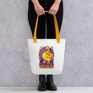 Könnte beinhalten: Weiße Tragetasche mit gelben Henkeln. Sie zeigt ein Cartoon-Huhn mit Sonnenbrille, Regenbogen und Sternen. Der Text "OWN YOUR WEIRD!" steht darunter. Die Tasche wird von einer Person getragen, die eine schwarze Hose und Stiefel trägt.