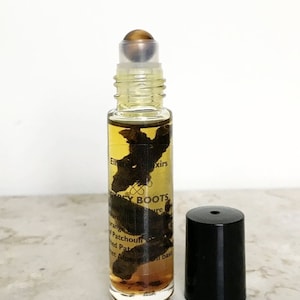 May include: A clear glass roller bottle with a black cap and a tiger's eye stone roller ball. The bottle contains a brown liquid and has a label that reads "Elixir Gypsy Boots Pure Essential Oil Blend". The label also lists the ingredients: "Orange, Patchouli, Dried Patchouli, Sweet Almond Oil, and Vanilla Bean."