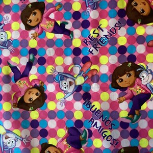 May include: Fabric featuring a repeating pattern of a cartoon girl and a monkey, set against a pink background with colorful polka dots. The words "Best Friends!" and "¡Buenos Amigos!" are also present.