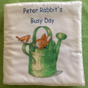May include: A white fabric book with the title "Peter Rabbit's Busy Day" printed in blue. The cover features an illustration of Peter Rabbit sitting in a green watering can with a red robin perched on the rim.