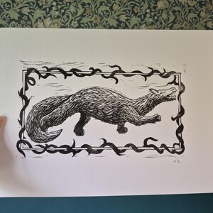 Peut inclure: Estampe en linogravure en noir et blanc représentant une créature ressemblant à un loup, encadrée d'une bordure décorative. L'animal est représenté en mouvement, la gueule ouverte. L'œuvre est sur papier blanc, tenue par une main, sur fond de papier peint floral et de mur bleu.