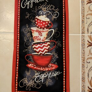 May include: A black and red kitchen towel with a stack of coffee cups and the words "Cappuccino, Latte, Espresso, Coffee" in white cursive writing.