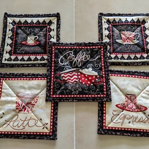 May include: Five black and white quilted pot holders with red and white accents. Each pot holder features a different coffee-themed design with a cup of coffee, latte, or espresso. The pot holders are decorated with the words "Coffee", "Espresso", and "Latte".