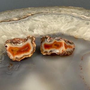 Puede incluir: Dos piedras de ágata pulidas con un llamativo patrón de bandas naranjas, rojas y blancas. Las piedras tienen un exterior áspero y marrón y un interior liso y brillante. Estas piedras preciosas naturales son ideales para la fabricación de joyas o coleccionar.