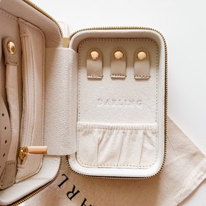 以下が含まれることがあります： 前面に「DARLING」という文字が刻印された、金色のアクセントが付いた白い革製のジュエリー旅行ケース。ケースが開いており、白い内装にはジュエリーを収納するための複数のコンパートメントがあります。
