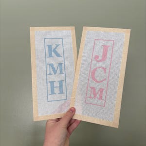 May include: Two rectangular needlepoint canvases with beige fabric borders. The left canvas features the blue letters KMH stacked vertically inside a blue rectangular frame. The right canvas features the pink letters JCM stacked vertically inside a pink frame.