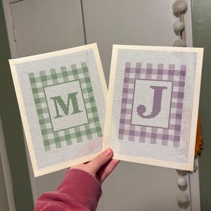May include: Two rectangular needlepoint canvases featuring a gingham check border pattern. One canvas displays a green letter M in the center, while the other canvas displays a purple letter J in the center. Both canvases have a white grid mesh.