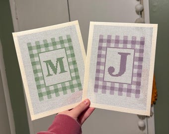 May include: Two rectangular needlepoint canvases featuring a gingham check border pattern. One canvas displays a green letter M in the center, while the other canvas displays a purple letter J in the center. Both canvases have a white grid mesh.