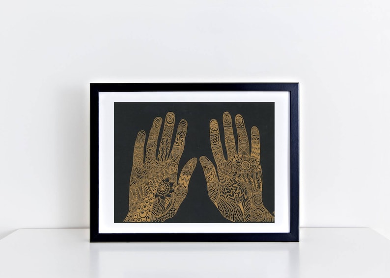 K&ouml;nnte beinhalten: Ein gerahmter Druck von zwei H&auml;nden mit aufwendigen goldenen Henna-Designs auf schwarzem Hintergrund.