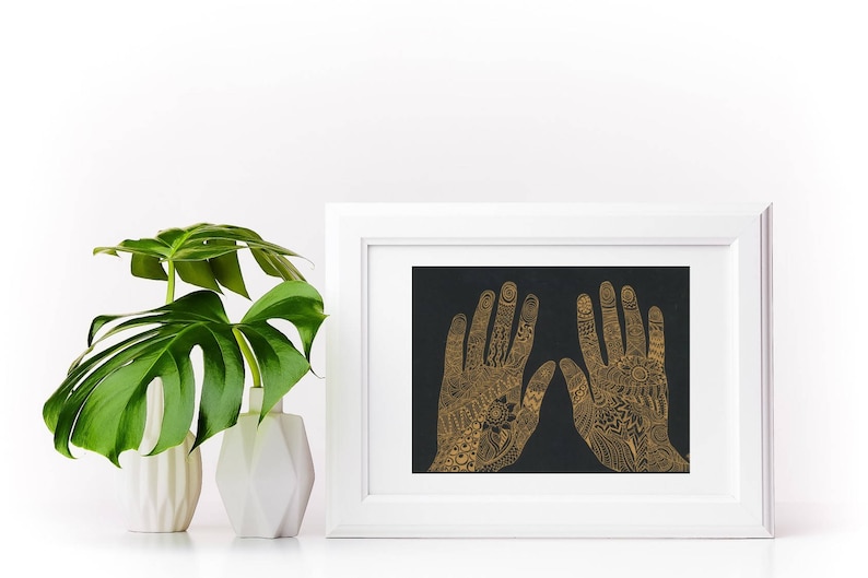 K&ouml;nnte beinhalten: Ein wei&szlig;er Rahmen mit schwarzem Hintergrund und zwei goldenen H&auml;nden mit komplizierten Designs. Die H&auml;nde sind erhoben, die Finger sind gespreizt.