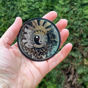 Könnte beinhalten: Ein runder, schwarzer Aufkleber mit einem goldenen und weißen Design. Das Design zeigt eine Sonne, Wellen, Wolken und ein Yin-Yang-Symbol.