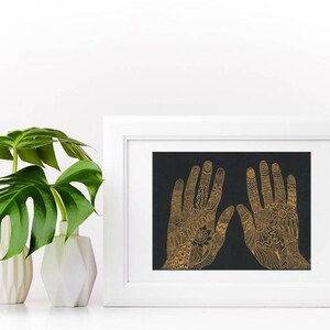K&ouml;nnte beinhalten: Ein gerahmter Druck von zwei goldenen H&auml;nden mit komplizierten Henna-Designs auf schwarzem Hintergrund.