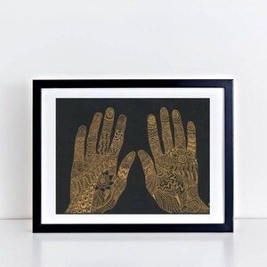 K&ouml;nnte beinhalten: Ein gerahmter Druck von zwei H&auml;nden mit aufwendigen goldenen Henna-Designs auf schwarzem Hintergrund.