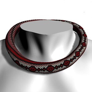 Op de afbeelding: Een rode, witte en zwarte kralenketting met een geometrisch patroon. De ketting is gemaakt van kleine, ronde kralen en wordt gedragen door een mannequin.
