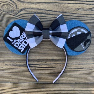 May include: A pair of denim mouse ears with a black and white checkered bow. One ear has a patch that says "I love the dark side" with a heart. The other ear has a black and gray Star Wars Death Star patch.