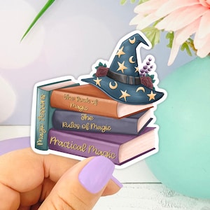 May include: A sticker featuring a stack of three books with titles like "The Rules of Magic" and "Practical Magic," topped with a blue witch's hat adorned with stars and a crescent moon. The sticker is held in a hand with purple nail polish.