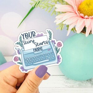 May include: A blue typewriter sticker with the text "YOUR story starts HERE" and floral accents. The sticker is held in a hand with purple nail polish.