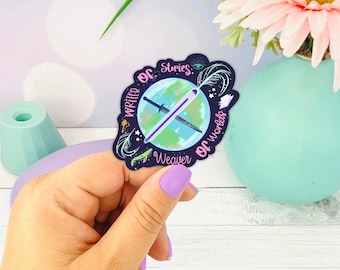 May include: A sticker with a dark blue border featuring a globe design with a sword and pen crossing it. The text "Writer of Stories, Weaver of Worlds" is written around the globe. The sticker is held by a person with purple nail polish.
