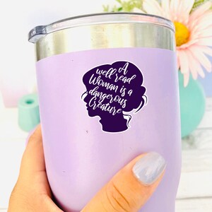 May include: A purple stainless steel tumbler with a white vinyl sticker that reads "A well read woman is a dangerous creature". The sticker features a silhouette of a woman's head.