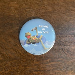 Op de afbeelding: Een ronde, lichtblauwe button met Kermit de Kikker die ontspant in een blauwe zwemband met een drankje. De button heeft de tekst "SUN DAYS ARE FUN DAYS!" en het copyright van Henson Associates, Inc.