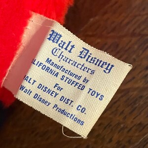 May include: Close-up of a vintage Walt Disney Characters tag. The tag is white with blue text, and reads "Walt Disney Characters" and "Manufactured by CALIFORNIA STUFFED TOYS For WALT DISNEY DIST. CO. Walt Disney Productions."