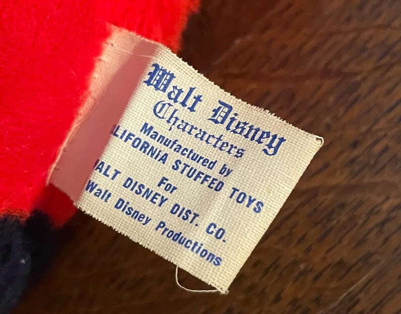 May include: Close-up of a vintage Walt Disney Characters tag. The tag is white with blue text, and reads "Walt Disney Characters" and "Manufactured by CALIFORNIA STUFFED TOYS For WALT DISNEY DIST. CO. Walt Disney Productions."