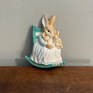 Puede incluir: Una decoración de pared de cerámica con un conejo marrón que sostiene dos conejos bebés en una mecedora verde.