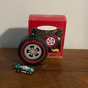 May include: A black and red Hallmark Keepsake ornament shaped like a Hot Wheels tire, with the words "Rally Case" on the side. A small green toy car is in front of the ornament. The ornament comes with a red box.