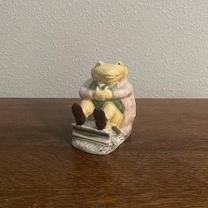 Op de afbeelding: Een keramieken beeldje van een kikker gekleed in een roze jas, groen vest en bruine schoenen, zittend op een stapel boeken. De kikker heeft een gele kop en lacht. Decoratief item.