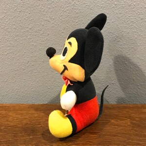 May include: A vintage Mickey Mouse soft toy. The toy has a black body, ears, and tail, with a yellow face, hands, and feet. Mickey wears red shorts and a red bow tie. The toy is sitting on a wooden surface.