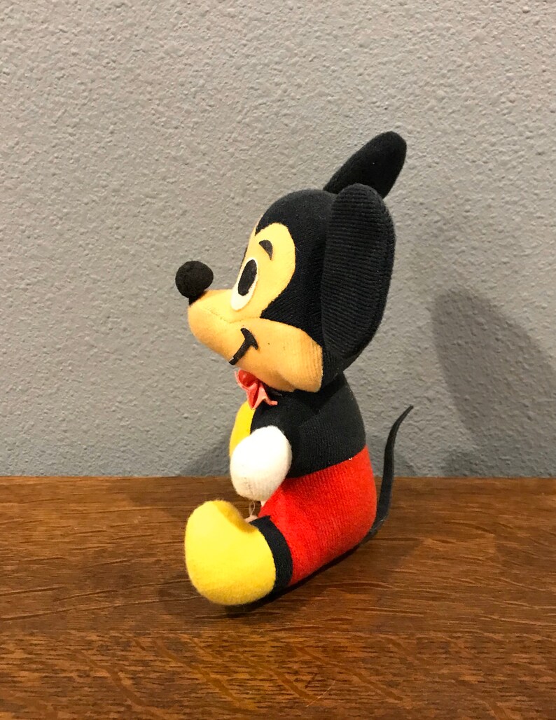 May include: A vintage Mickey Mouse soft toy. The toy has a black body, ears, and tail, with a yellow face, hands, and feet. Mickey wears red shorts and a red bow tie. The toy is sitting on a wooden surface.