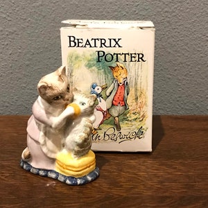 Puede incluir: Una figurita de Beatrix Potter de un gato con un vestido rosa alimentando a un gatito. La figurita está sentada sobre una base amarilla y azul. La figurita está empaquetada en una caja con el logotipo de Beatrix Potter y el título "The Tale of Mrs. Tiggy-Winkle".